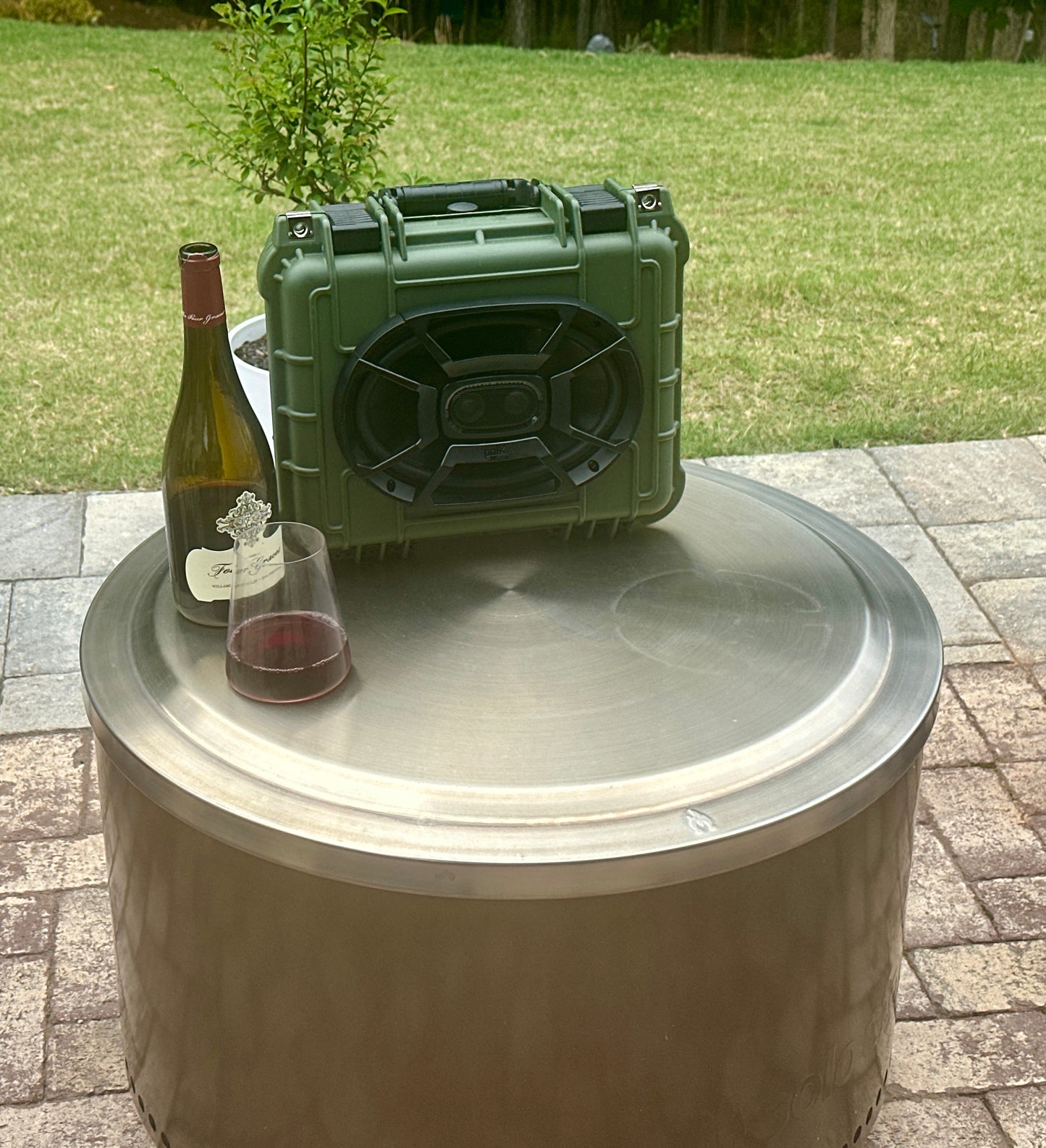 Green portable speaker on a metal fire pit with a bottle of wine and glass.